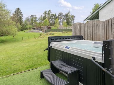 An outdoor hot tub with steps next to a wooden fence and a large grassy area with trees and a wooden cabin in the background at Maple - Woodland Cottages in Bowness-on-Windermere