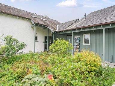 An exterior view of a building with green and white walls surrounded by plants and shrubs at Maple - Woodland Cottages in Bowness-on-Windermere