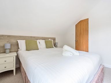 A bedroom with a large bed with white linens and green pillows a wooden headboard a wooden door and a white nightstand with a lamp at Maple - Woodland Cottages in Bowness-on-Windermere
