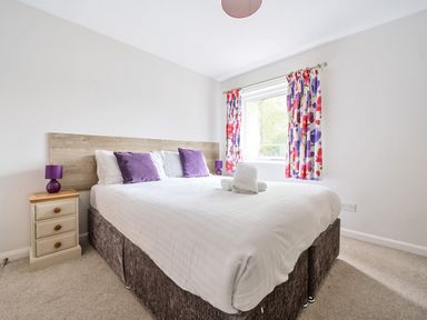 A bedroom with a double bed purple cushions floral curtains and bedside drawers at Maple - Woodland Cottages in Bowness-on-Windermere