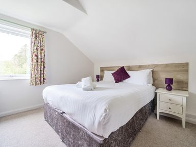 A bedroom with a double bed white bedding purple cushion and lamp next to a window with floral curtains at Maple - Woodland Cottages in Bowness-on-Windermere