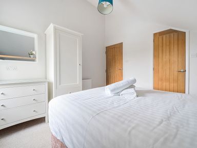 A bedroom with a bed with white linens and folded towels a white dresser with a mirror and a plant a white wardrobe and two wooden doors Maple - Woodland Cottages Bowness-on-Windermere