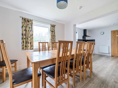 A dining area with a wooden table and eight chairs next to a window with patterned curtains at Maple - Woodland Cottages in Bowness-on-Windermere