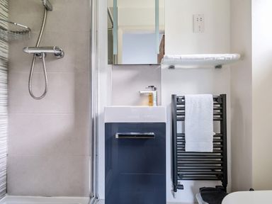 A bathroom with a shower unit a small sink with cabinet a mirror a towel rack with a white towel and a shelf with folded towels at Maple - Woodland Cottages in Bowness-on-Windermere