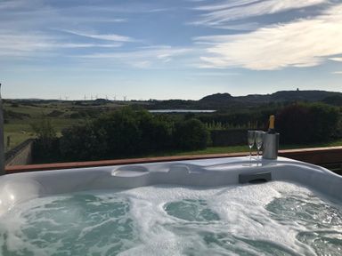 A hot tub with a bottle in an ice bucket and two glasses on a wooden railing overlooking a countryside landscape with hills and wind turbines at Rhos Helyg in Penysarn near Amlwch
