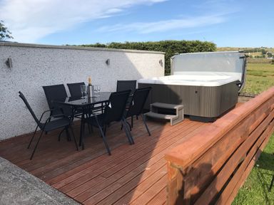 An outdoor wooden deck with a black dining table and chairs a hot tub and a wooden fence at Rhos Helyg in Penysarn near Amlwch