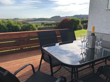 An outdoor wooden deck with a glass table and black chairs a bottle in a cooler with glasses and greenery and hills in the background at Rhos Helyg in Penysarn near Amlwch