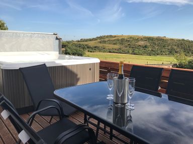 An outdoor patio with a hot tub a glass table with a champagne bottle and four glasses and black chairs at Rhos Helyg in Penysarn near Amlwch