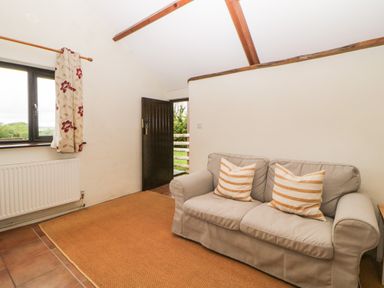 A room with a beige sofa and two striped cushions near a window and an open door at Nestling Barn in Peter Tavy near Tavistock