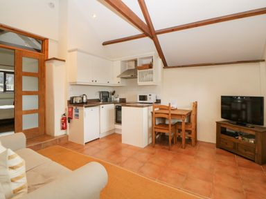 A kitchen area with white cabinets wooden dining table and chairs and a television on a wooden stand at Nestling Barn in Peter Tavy near Tavistock