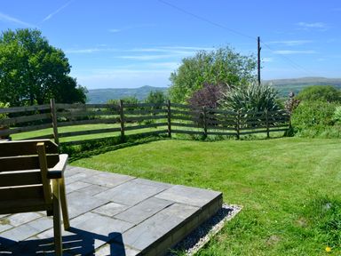 A garden with a wooden bench on a stone patio and a wooden fence with bushes and trees at Nestling Barn Peter Tavy near Tavistock