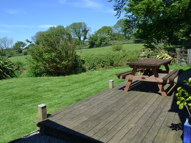 A wooden picnic table on a deck beside a lawn with bushes and trees at Nestling Barn in Peter Tavy near Tavistock