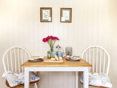 A small wooden table with two white chairs a vase with flowers a cutting board with scones and plates with jam and cream on a wooden wall at Bramble Burlawn near Wadebridge