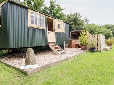 A green metal shepherds hut with cream door and windows on a gravel platform with potted plants and wooden fenced area at Bramble Burlawn near Wadebridge