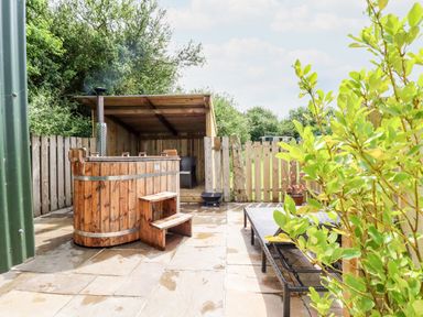 An outdoor patio area with a wooden hot tub stairs lounge chairs and greenery at Bramble Burlawn near Wadebridge