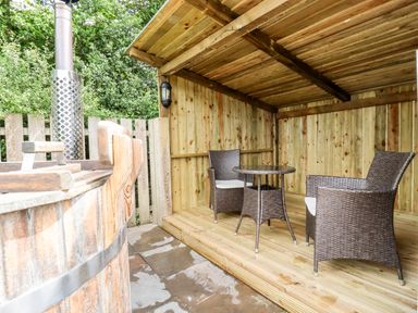 A wooden deck with two woven chairs and a small round table under a wooden shelter next to a wooden hot tub at Bramble Burlawn near Wadebridge