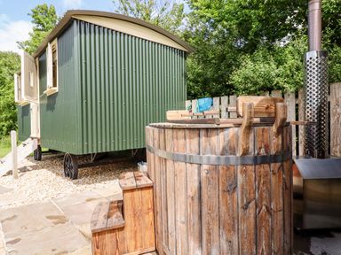 An outdoor area with a green corrugated metal shepherds hut and a wooden hot tub with metal bands near a metal chimney at Bramble Burlawn near Wadebridge