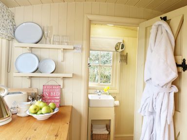 A kitchen counter with a bowl of green apples plates glasses and a sign next to a doorway with a white bathrobe hanging and a small bathroom with a sink and window at Willow Burlawn near Wadebridge