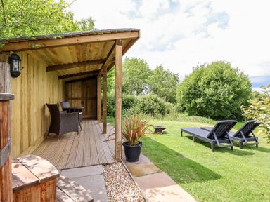 A wooden covered patio with chairs and table next to a grassy garden with two lounge chairs at Willow Burlawn near Wadebridge