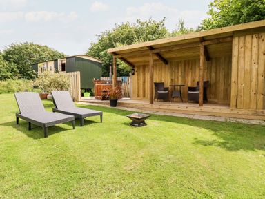 A grassy backyard with two lounge chairs a fire pit a wooden covered seating area with two chairs and a table and a green caravan at Willow Burlawn near Wadebridge