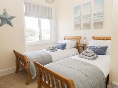 A bedroom with two single wooden beds, gray blankets, pillows with text, a window with striped blinds, and three seaside paintings at Oak Lodge in East Runton