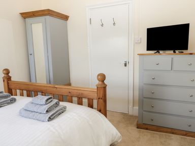 A bedroom with a wooden bed frame with towels on the bed a grey wardrobe with a mirror a white door and a grey chest of drawers with a television on top at Oak Lodge in East Runton