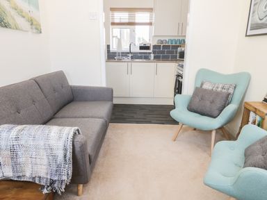 A small living room with a gray sofa and two light blue armchairs facing a kitchen with white cabinets and a window at Oak Lodge in East Runton