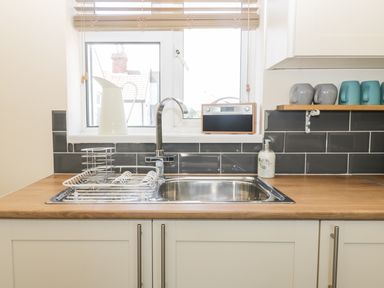 A kitchen sink with a drying rack on a wooden countertop below a window with blinds and a white pitcher and radio on the windowsill at Oak Lodge in East Runton