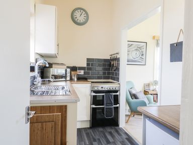 A kitchen with a sink, microwave, oven, and clock on the wall leading to a room with an armchair at Oak Lodge in East Runton