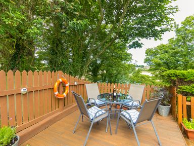 An outdoor wooden deck with a round glass table surrounded by four chairs with cushions and a wooden fence with a life buoy at Estuary View in Warkworth