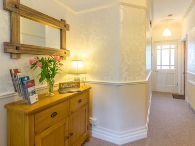 A hallway with a wooden cabinet holding a lamp flowers and a welcome sign at Estuary View in Warkworth