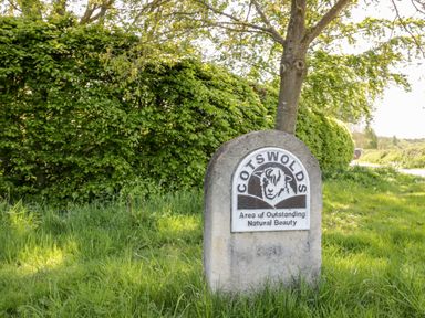 A stone sign with a sheep illustration and text about Cotswolds area in a grassy area near trees at Beechnut Cottage in Tewkesbury