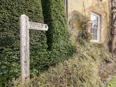 A wooden Cotswold Way public footpath signpost near a hedge and a stone building with a window at Beechnut Cottage in Tewkesbury
