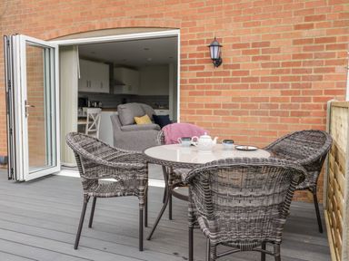 An outdoor patio with a round table and four wicker chairs near an open door leading to a living room with a grey armchair at Beechnut Cottage in Tewkesbury