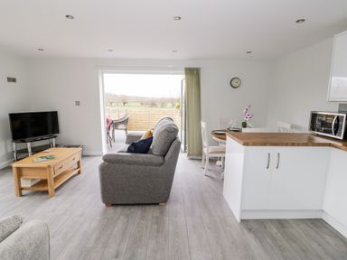 A living room with a gray sofa a wooden coffee table and a TV next to a kitchen with white cabinets and a microwave at Beechnut Cottage in Tewkesbury