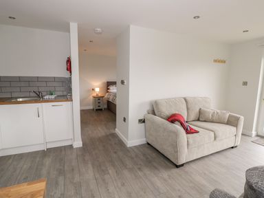 A living area with a beige sofa and blanket near a kitchen area and a bedroom with a lamp at Beechnut Cottage in Tewkesbury