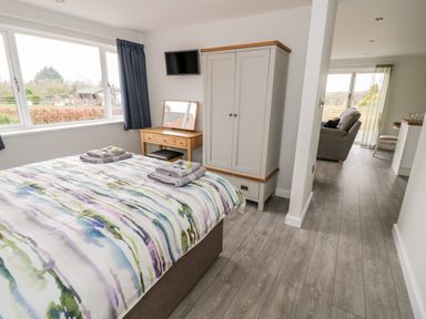 A bedroom with a bed covered with a patterned duvet next to a window a wardrobe a small wooden desk with a mirror and a television on the wall at Beechnut Cottage in Tewkesbury