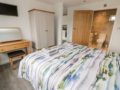 A bedroom with a bed with colorful striped bedding a wooden wardrobe a dressing table with a mirror a mounted TV and an open door leading to a bathroom at Beechnut Cottage in Tewkesbury