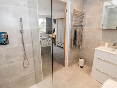 A bathroom with a shower, towel rack, sink, and trash bin with a view of a bedroom through an open door at Beechnut Cottage in Tewkesbury