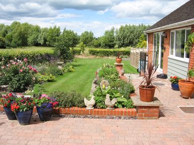 A garden with flower pots on a brick patio and a lawn bordered by flower beds at Beechnut Cottage in Tewkesbury
