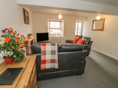 A living room with black leather sofas a wooden cabinet with flowers a small TV and a window at Brickfield Cottage in Machynlleth