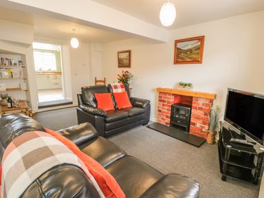 A living room with black leather sofas with orange cushions a fireplace and a television at Brickfield Cottage in Machynlleth