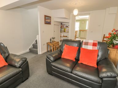 A living room with black leather sofas with red cushions a small wooden table with a bottle and glasses stairs and a view through a doorway to a kitchen at Brickfield Cottage in Machynlleth