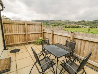 A small outdoor patio with a black glass table and four black chairs enclosed by wooden fences with a view of green fields and hills at Brickfield Cottage in Machynlleth