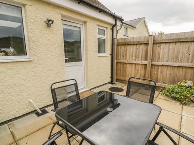 A patio area with a glass table and four chairs outside a light yellow building with a wooden fence and some plants at Brickfield Cottage in Machynlleth