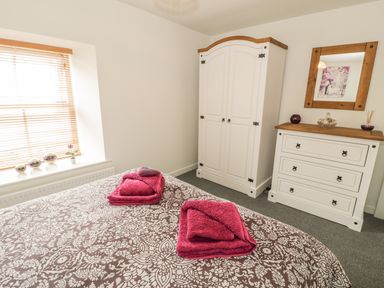 A bedroom with a bed and folded magenta towels on it a window with blinds white wardrobe and dresser with a mirror at Brickfield Cottage in Machynlleth