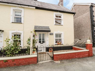The front of a cream colored cottage with white framed windows a black metal gate and flower pots at Brickfield Cottage in Machynlleth