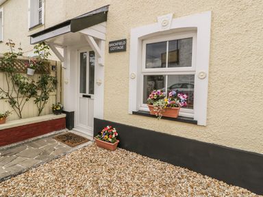 The entrance of Brickfield Cottage with a white door under a small overhang and flower pots on gravel and windowsill in Machynlleth