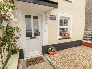 Exterior view of a cottage entrance with a white door, windowsill flowers, and a doormat at Brickfield Cottage in Machynlleth