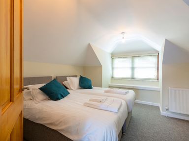 A bedroom with two single beds with white bedding and teal pillows under a sloped ceiling near a window with blinds at Rocky Close in St Ives